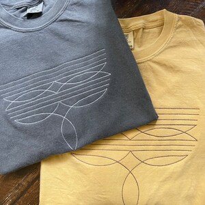 Puede incluir: Dos camisetas con dise&ntilde;os geom&eacute;tricos. La camiseta de arriba es gris oscuro con un dise&ntilde;o bordado blanco. La camiseta de abajo es amarilla con un dise&ntilde;o bordado marr&oacute;n.