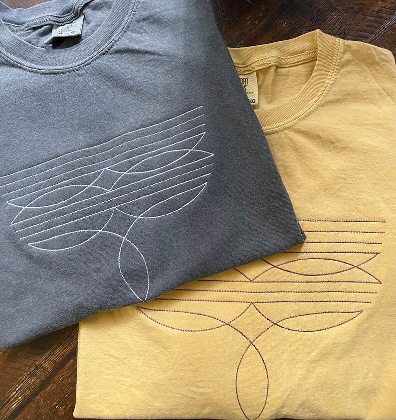 Puede incluir: Dos camisetas con dise&ntilde;os geom&eacute;tricos. La camiseta de arriba es gris oscuro con un dise&ntilde;o bordado blanco. La camiseta de abajo es amarilla con un dise&ntilde;o bordado marr&oacute;n.