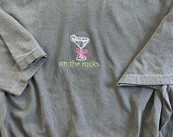 May include: A gray t-shirt with a white embroidered image of a cocktail glass with a pink ribbon and the text "on the rocks."