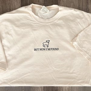 K&ouml;nnte beinhalten: Ein wei&szlig;es T-Shirt mit einer schwarzen Umrandung eines Schafes und dem Text "BUT NOW I'M FOUND".