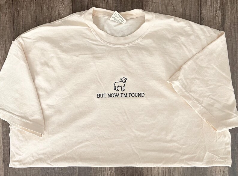 K&ouml;nnte beinhalten: Ein wei&szlig;es T-Shirt mit einer schwarzen Umrandung eines Schafes und dem Text "BUT NOW I'M FOUND".