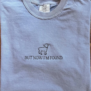 K&ouml;nnte beinhalten: Ein hellblaues T-Shirt mit einem kleinen, schwarzen gestickten Lamm und dem Text "BUT NOW I'M FOUND".
