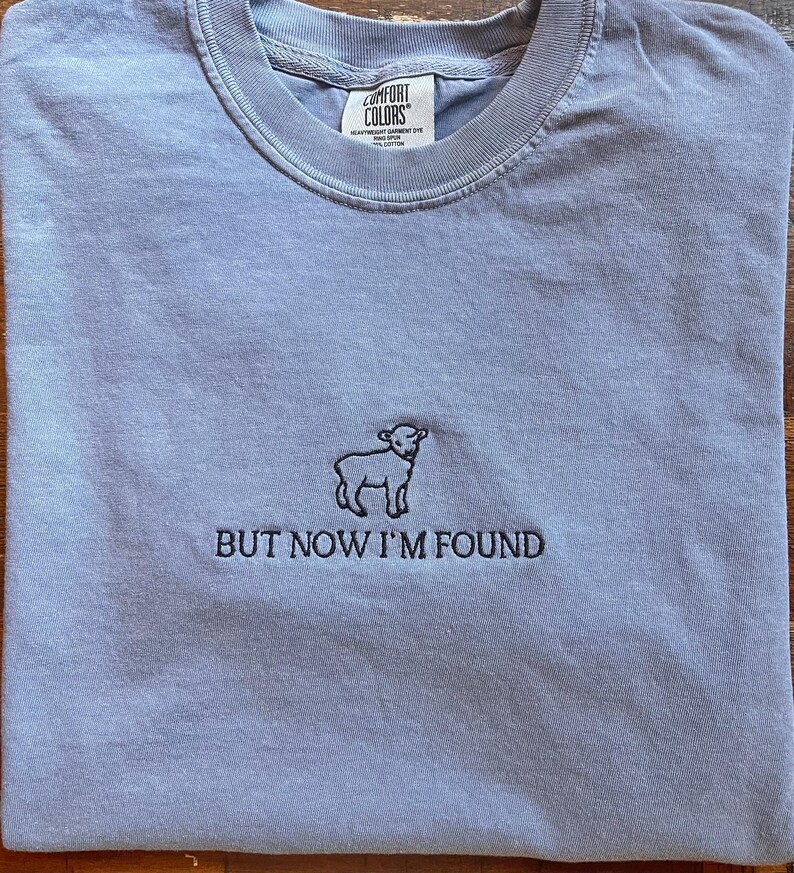 K&ouml;nnte beinhalten: Ein hellblaues T-Shirt mit einem kleinen, schwarzen gestickten Lamm und dem Text "BUT NOW I'M FOUND".