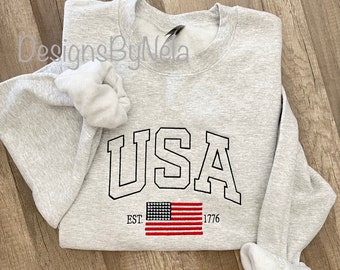 Puede incluir: Sudadera gris con un gráfico USA negro y una bandera estadounidense con el texto "EST. 1776".