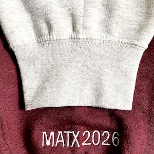 May include: Close-up of two sweatshirts. The top sweatshirt is light gray with "Class of 2026" embroidered in brown thread. The bottom sweatshirt is maroon with "MATX 2026" embroidered in white thread. The sweatshirts are likely for a school or class.