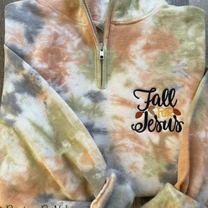 May include: A tie-dye quarter zip sweatshirt with the text "Fall for Jesus" embroidered on the front. The sweatshirt is in shades of brown, green, and gray.