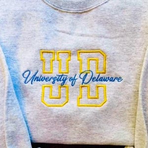 May include: Three sweatshirts with embroidered university logos. The first is white with "IU" and "Indiana University" in red. The second is gray with "UD" and "University of Delaware" in yellow and blue. The third is black with "MSU" and "Michigan State University" in green.