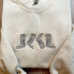 Puede incluir: Una sudadera beige con un diseño bordado en negro de cuatro botas de vaquero. Las botas están dispuestas en fila, con las puntas apuntando hacia la derecha.