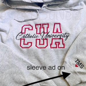 May include: A light gray hooded sweatshirt with the embroidered text "CUA Catholic University" in red and black. The sleeve has the initials "TMS" and a red heart.