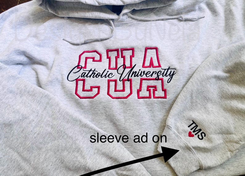 May include: A light gray hooded sweatshirt with the embroidered text "CUA Catholic University" in red and black. The sleeve has the initials "TMS" and a red heart.