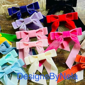 May include: A collection of velvet bows in various colors, including blue, green, yellow, pink, and red. The bows have long, flowing tails and are arranged on a wooden surface. The text "Designs By Nela" is visible.