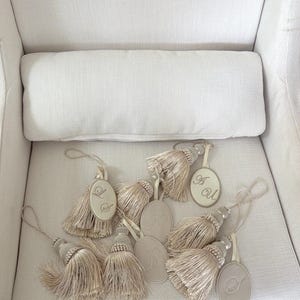 May include: A collection of beige decorative tassels, each featuring an oval-shaped tag with an embroidered monogram letter. The tassels are made of fine, light-colored thread and include a loop at the top for hanging, arranged in a cluster.