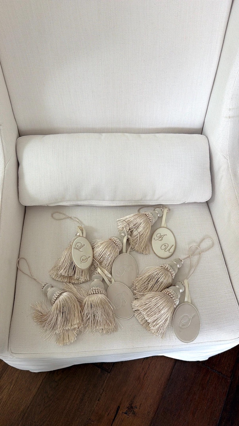 May include: A collection of beige decorative tassels, each featuring an oval-shaped tag with an embroidered monogram letter. The tassels are made of fine, light-colored thread and include a loop at the top for hanging, arranged in a cluster.