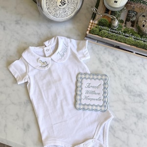 Op de afbeelding: Witte babybody met een Peter Pan kraag. De kraag is geborduurd met de naam "Israel" en een schommelpaard. Een bijpassend naamlabel met de naam "Israel William Harper" is ook inbegrepen.