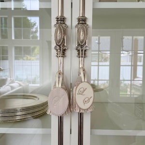 May include: Two decorative tassels with oval fabric tags hanging from silver-toned metal cabinet handles. One tag features an embroidered letter S, while the other displays the intertwined letters L and S. The tassels are light beige with fringe.