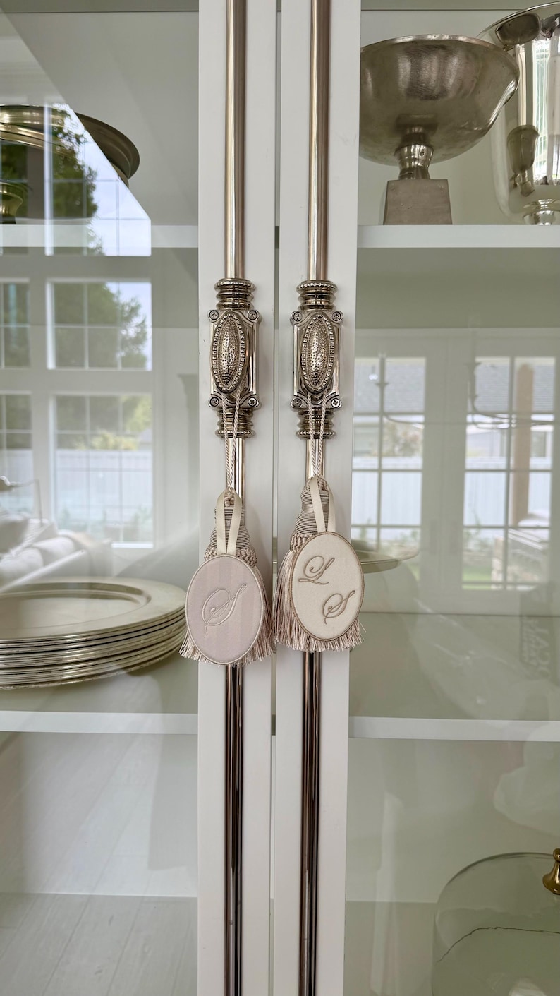 May include: Two decorative tassels with oval fabric tags hanging from silver-toned metal cabinet handles. One tag features an embroidered letter S, while the other displays the intertwined letters L and S. The tassels are light beige with fringe.