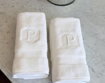 May include: Two folded white hand towels featuring an embroidered letter P within a square border, positioned next to a cylindrical candle with a metallic silver mercury glass finish and a decorative silver lid with intricate circular patterns.