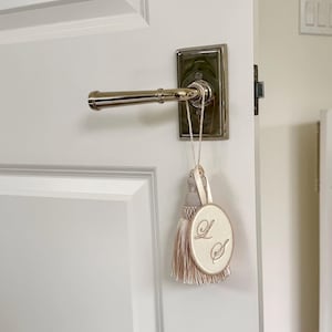 May include: A beige oval fabric ornament with the embroidered letters L and S, featuring a matching beige tassel and a thin cord loop. The ornament hangs from a polished silver-toned metal door handle mounted on a white paneled door.