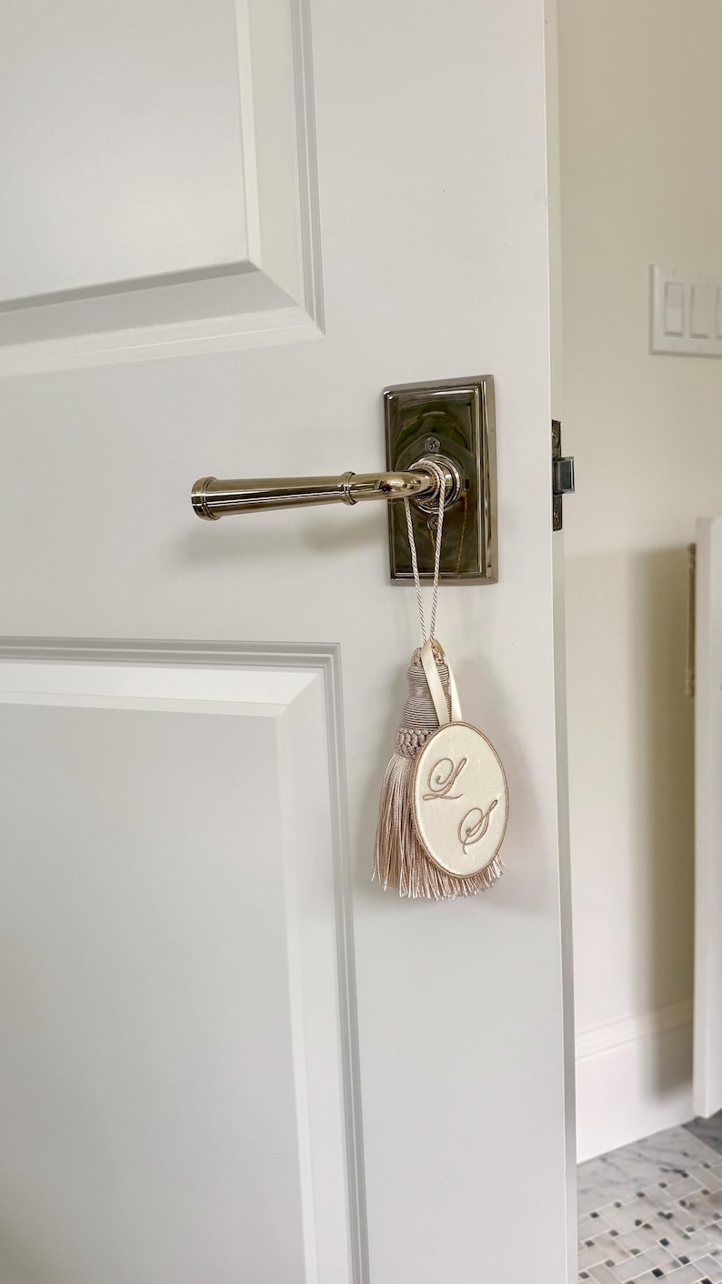 May include: A beige oval fabric ornament with the embroidered letters L and S, featuring a matching beige tassel and a thin cord loop. The ornament hangs from a polished silver-toned metal door handle mounted on a white paneled door.