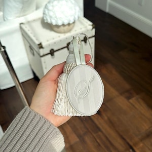 May include: An oval-shaped off-white fabric ornament featuring a subtle vertical striped pattern and an embroidered script letter S in the center. The ornament is attached to a matching off-white decorative tassel with a braided cord loop for hanging.