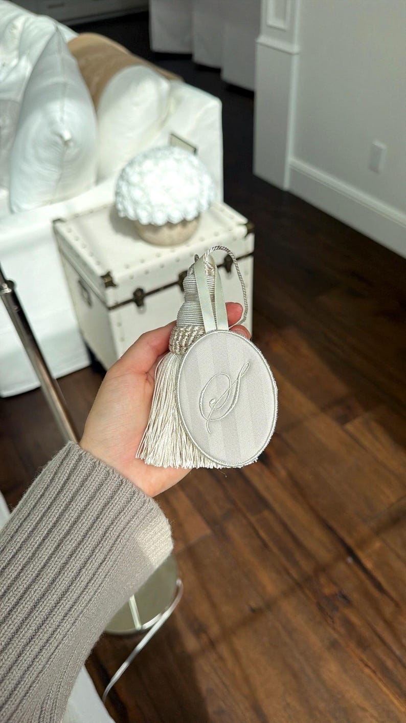 May include: An oval-shaped off-white fabric ornament featuring a subtle vertical striped pattern and an embroidered script letter S in the center. The ornament is attached to a matching off-white decorative tassel with a braided cord loop for hanging.