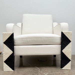 Pode incluir: Uma poltrona branca com um design geométrico nos apoios de braços. Os apoios de braços apresentam um padrão tecido em preto e creme. A cadeira tem um design simples e moderno e está sobre um fundo branco liso.