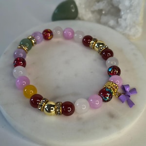 May include: A multi-colored beaded bracelet with a purple bow charm. The bracelet features pink, white, yellow, green, and red beads with gold accents.