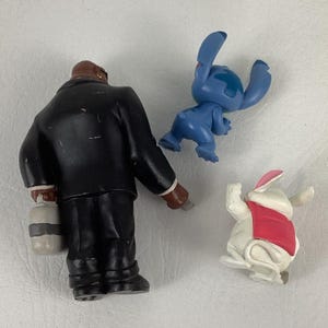 May include: Three small figurines: a man in a black suit holding a container, a blue alien-like character, and a white rabbit wearing a red vest. The figurines are made of plastic and have painted details.