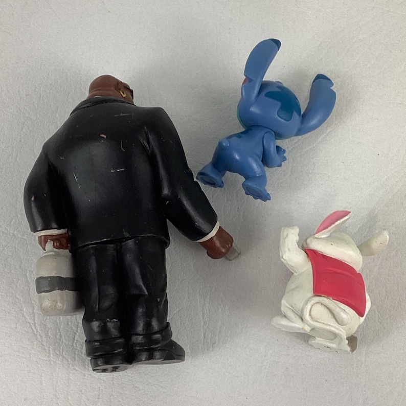May include: Three small figurines: a man in a black suit holding a container, a blue alien-like character, and a white rabbit wearing a red vest. The figurines are made of plastic and have painted details.