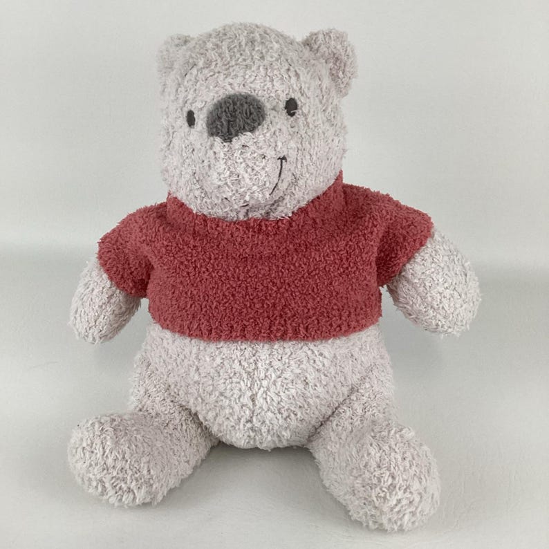 May include: A plush Winnie the Pooh stuffed animal. The bear has a gray body, a dark red shirt, and a gray nose. The bear is sitting with its arms and legs outstretched.