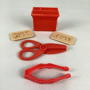 May include: A red plastic toy medical kit set including a rectangular storage container, a pair of red scissors, red tweezers, and two beige plastic bandages with heart-shaped centres. The container features a moulded logo on the front side.