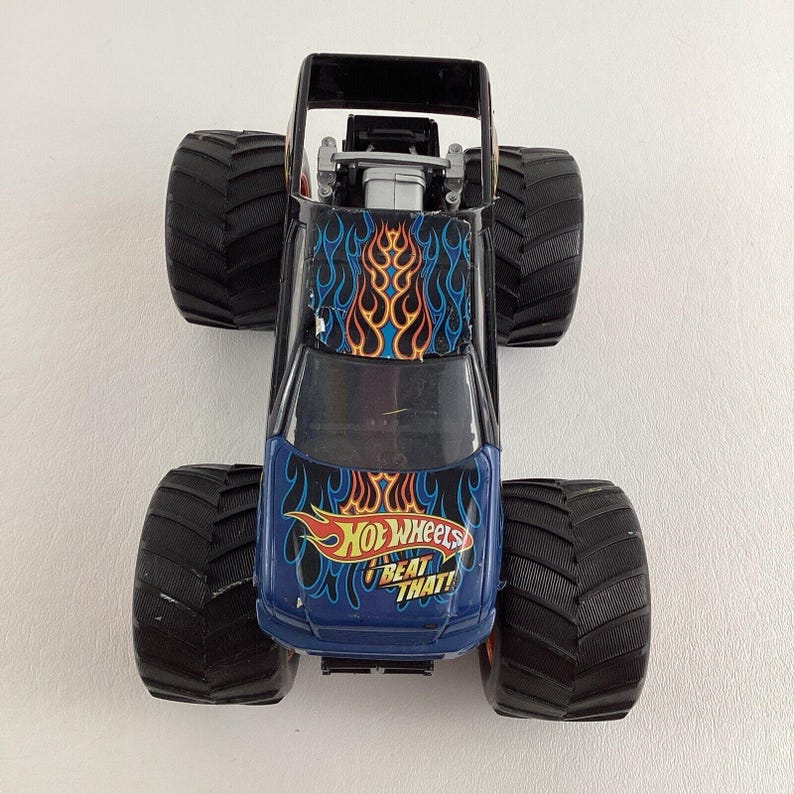Puede incluir: Un juguete de cami&oacute;n monstruo Hot Wheels negro y azul con neum&aacute;ticos negros grandes. El cami&oacute;n presenta un dise&ntilde;o de llamas en el cap&oacute; y el texto "Hot Wheels Beat That!"