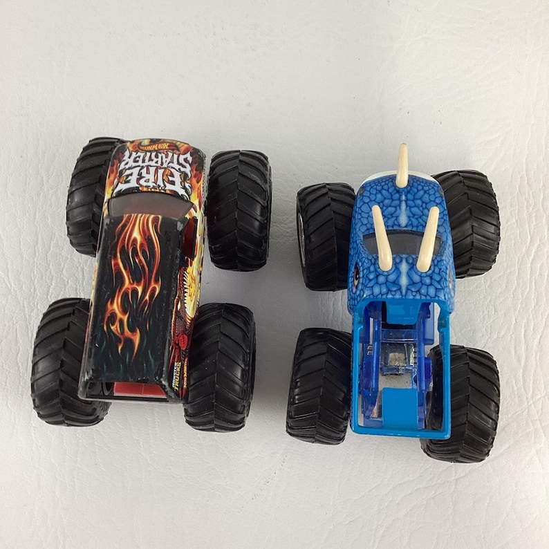 May include: Two toy monster trucks. One is black with red and yellow flames and the text "Fire Starter". The other is blue with a dinosaur head design. Both have large black tires.