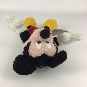 May include: A plush Mickey Mouse toy with a red bow, black ears, and a black nose. The toy has white gloves and yellow shoes. The face is pink and the toy is lying on its back.