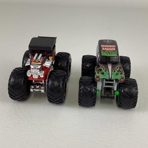 May include: Two toy monster trucks. One is red with a skull design and black tires. The other is green with a flame design and the text "Grave Digger" on top. Both have large black tires.