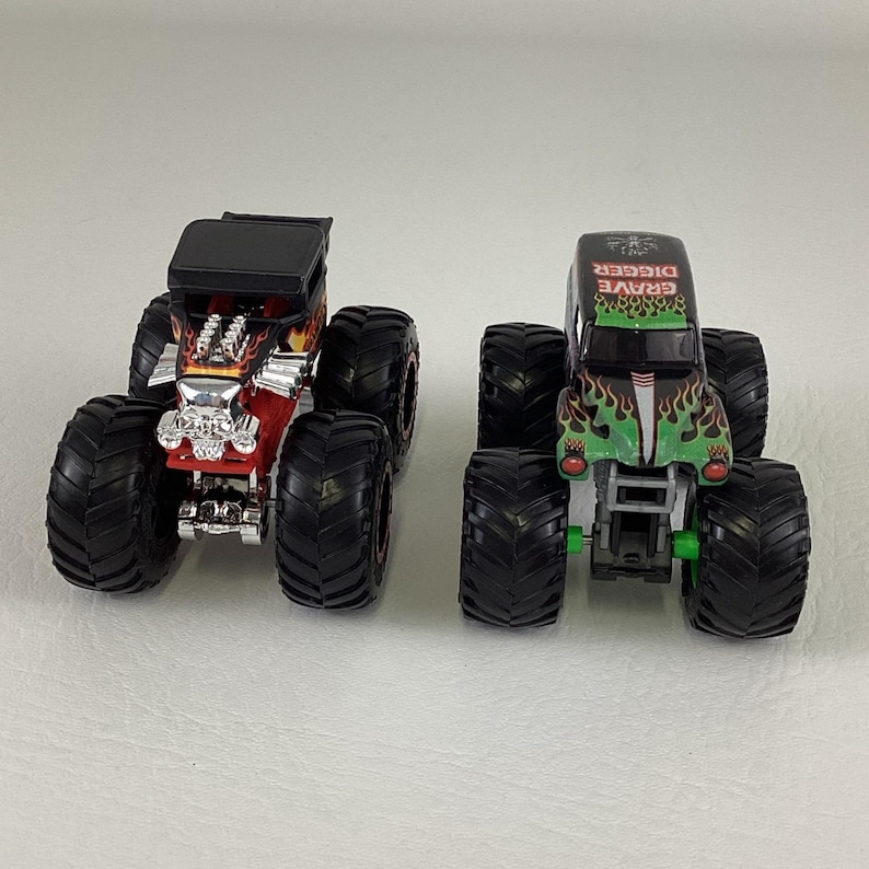 May include: Two toy monster trucks. One is red with a skull design and black tires. The other is green with a flame design and the text "Grave Digger" on top. Both have large black tires.