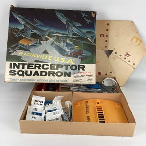 Può includere: Kit di gioco vintage Plasticville U.S.A. Interceptor Squadron. La scatola presenta illustrazioni di aerei a reazione e una scena aeroportuale. Il kit include vari componenti in plastica, edifici, aerei e accessori, contenuti in una scatola di cartone.