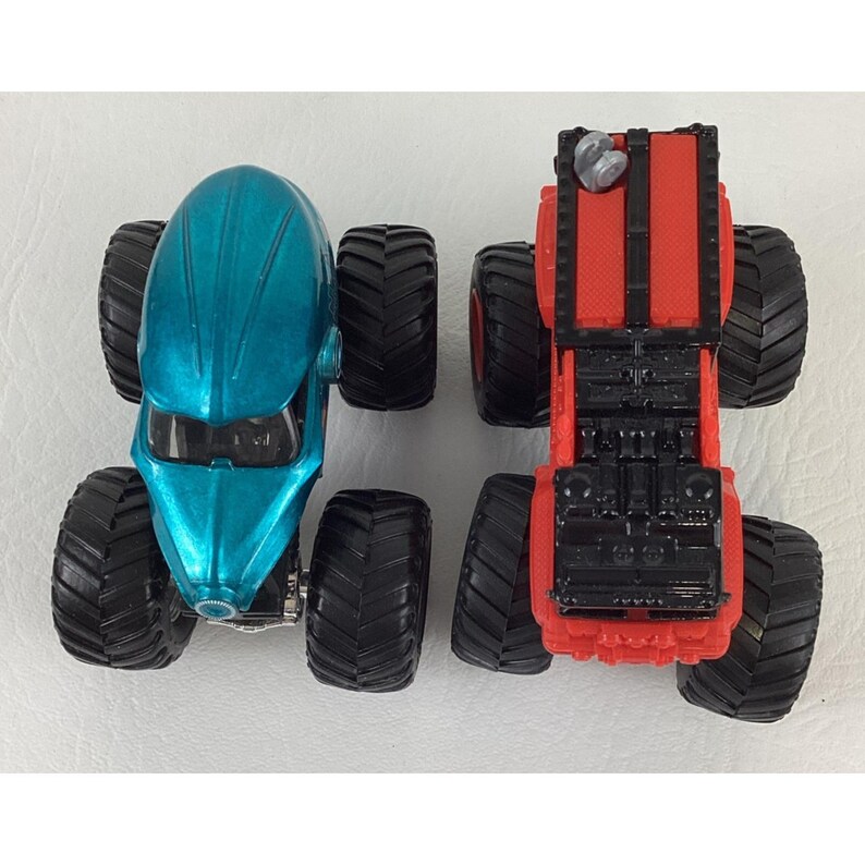 Pu&ograve; includere: Due monster truck giocattolo. Uno &egrave; verde acqua con una cabina arrotondata, l'altro &egrave; rosso con una cabina rettangolare. Entrambi hanno grandi pneumatici neri. I camion sono visti dall'alto.