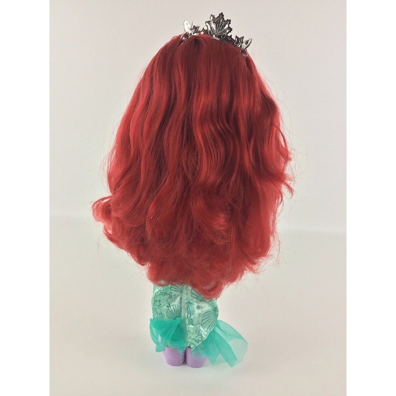 May include: A red-haired doll wearing a green and blue dress with a silver crown. The doll is standing on a white surface.