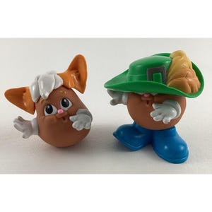 May include: Two small brown potato-shaped figures with painted faces. One figure is wearing a green hat with a feather and blue shoes. The other figure has white fur and orange ears.