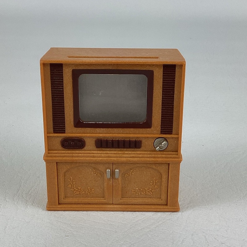 Op de afbeelding: Een miniatuurtelevisie in vintage-stijl in bruine tinten. De tv heeft een rechthoekig scherm, een bedieningspaneel en een kast met deuren. Het ontwerp omvat decoratieve details en een knop, die een retro-esthetiek oproepen.