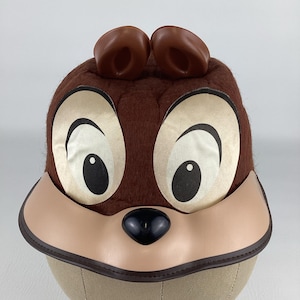 Peut inclure: Un chapeau Chip marron avec de grands yeux de dessin animé, un museau beige et un nez noir. Le chapeau a deux protubérances marron en forme d'oreille sur le dessus. Le chapeau est conçu pour ressembler au personnage Disney Chip.