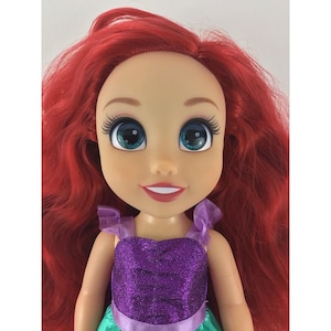 May include: A red-haired doll with blue eyes wearing a purple and green dress. The doll has a smiling face and is looking directly at the camera.