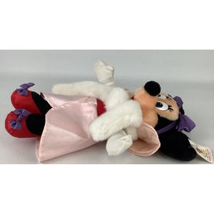 May include: A plush Minnie Mouse doll lying on its side, wearing a light pink satin dress, a white faux fur stole, red shoes with purple bows, and a matching purple bow on its head. The doll has black ears, white gloves, and a tag attached.