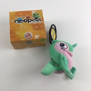 May include: A green and pink Neopets plush toy with a Burger King logo on the box. The toy is attached to a keychain with a black ring.