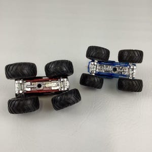 May include: Two toy monster trucks, one red and one blue, are shown from the underside. The trucks have large black tires and silver chassis details. The vehicles are positioned on a light gray surface.