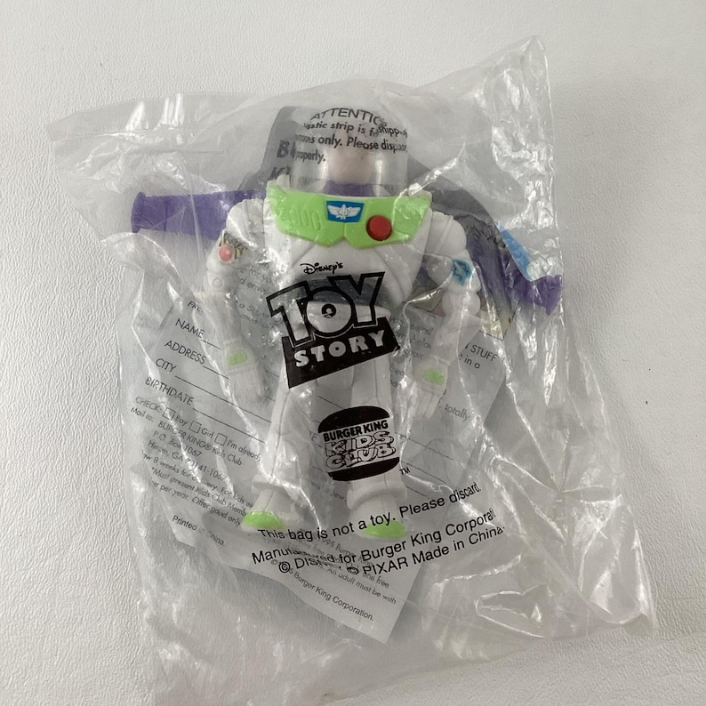 May include: A Buzz Lightyear action figure from the Disney Pixar Toy Story franchise, packaged in a clear plastic bag. The figure is white with green and purple accents, and the packaging includes the Burger King Kids Club logo.