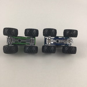 May include: Two toy monster trucks, one green and one blue, with black wheels. The trucks are shown from the bottom, revealing the chassis and axles.
