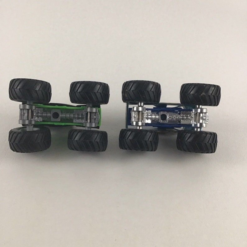 May include: Two toy monster trucks, one green and one blue, with black wheels. The trucks are shown from the bottom, revealing the chassis and axles.