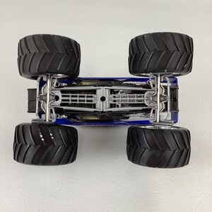 Op de afbeelding: Een blauw monstertruckspeelgoed met vier grote zwarte banden. De onderkant van de truck is zichtbaar en toont het chassis en de ophangingsdetails. De banden hebben een diep profiel.